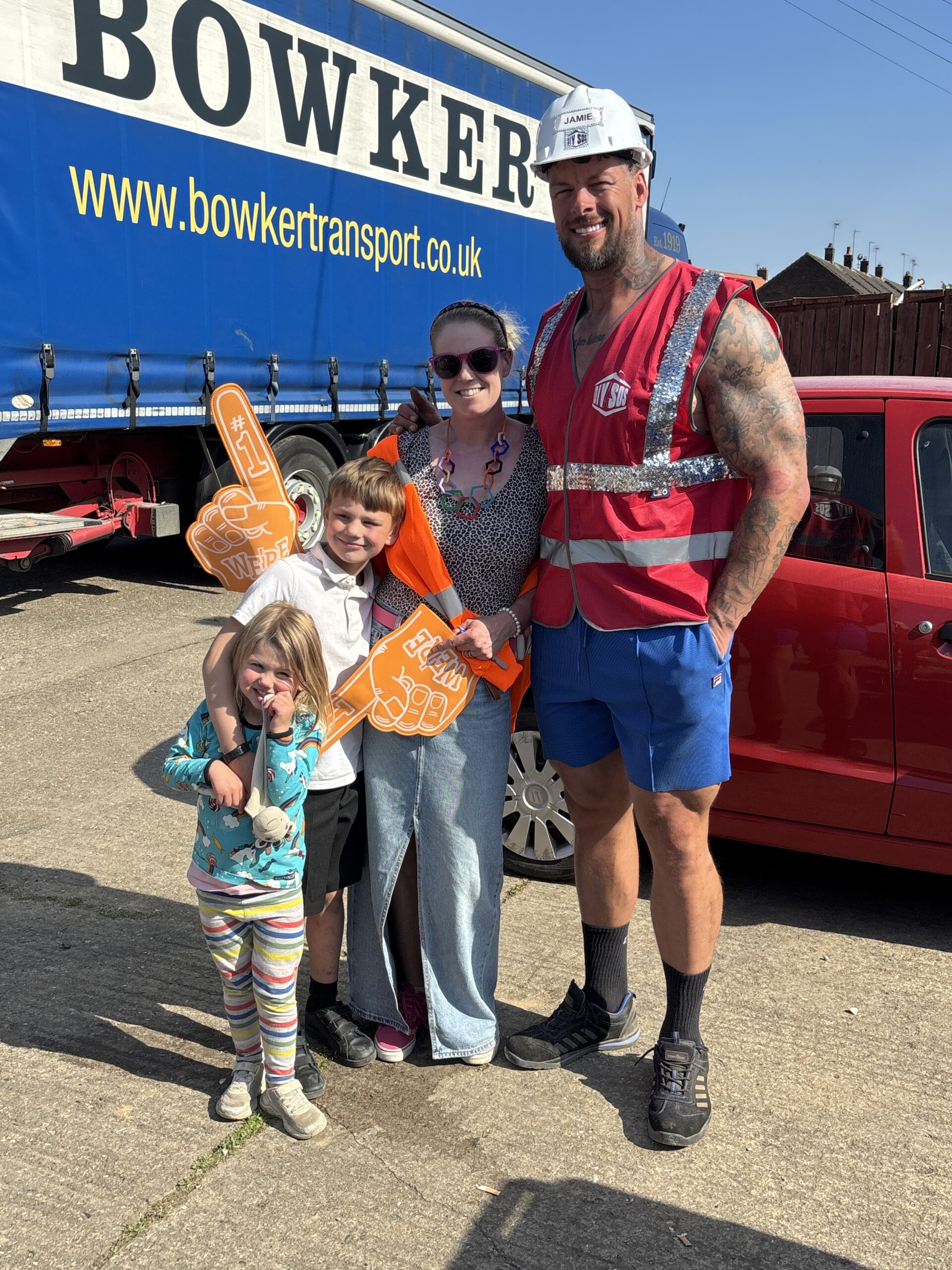 A group standing in a sunny outdoor lot in front of a blue truck and a red car. A man in construction gear and a hard hat poses with a woman and two children. The group is smiling, and the boy is holding up a large orange foam hand.
