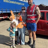 A group photo taken outdoors in front of a large blue transport truck labeled "BOWKER." The group includes a man wearing a white hard hat labeled "Jamie," a red high-visibility vest, and blue shorts. He stands next to a smiling woman in sunglasses and a leopard-print top, and two young children. The boy holds an orange foam finger, and the girl holds a stuffed animal.