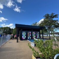A modern, black modular building with large windows and colorful square artwork panels on the side. In the foreground, there is an outdoor ping pong table on a paved surface. To the left, a crowd of people, some wearing high-visibility vests, stands behind a white metal fence.