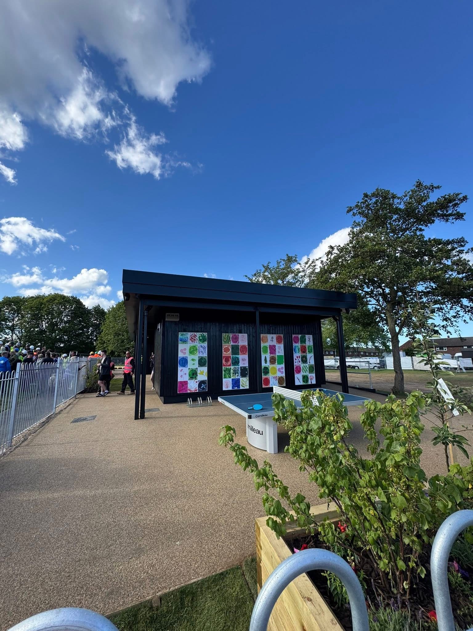 A modern, black modular building with large windows and colorful square artwork panels on the side. In the foreground, there is an outdoor ping pong table on a paved surface. To the left, a crowd of people, some wearing high-visibility vests, stands behind a white metal fence.