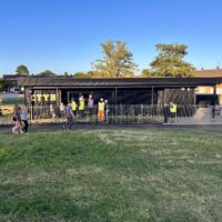 A wide landscape shot of a black, single-story building with the gold letters "CTYH" attached to the exterior wall. The building is surrounded by temporary metal fencing, and several people in high-visibility vests are walking near the entrance. The foreground features a grassy slope.