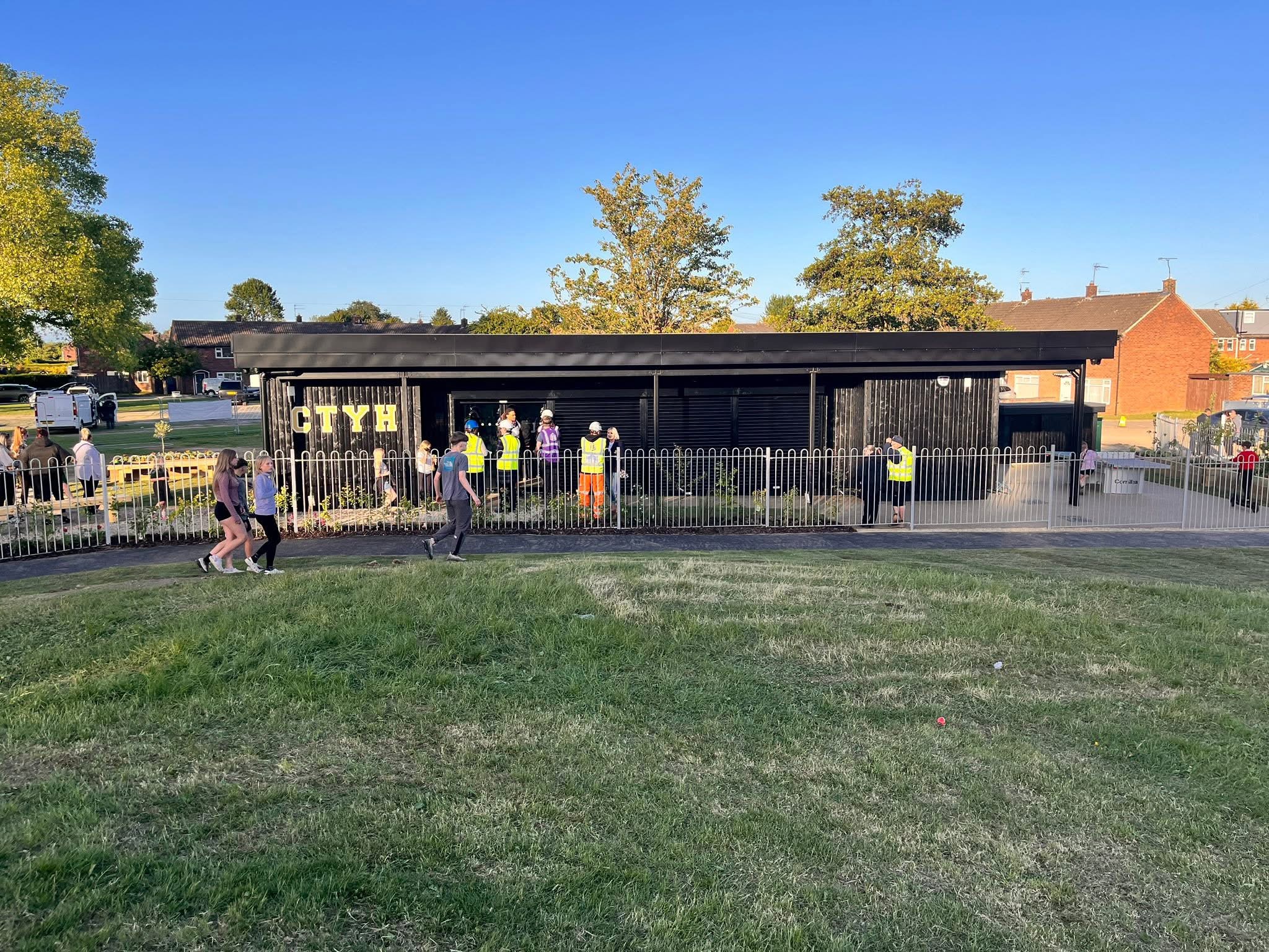A wide landscape shot of a black, single-story building with the gold letters "CTYH" attached to the exterior wall. The building is surrounded by temporary metal fencing, and several people in high-visibility vests are walking near the entrance. The foreground features a grassy slope.