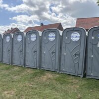 A close-up view of a row of grey portable toilets situated on grass. The toilet on the far right has a handwritten paper sign taped to the door that reads "WOMEN ONLY." The tops of brick houses and leafy trees are visible in the background.