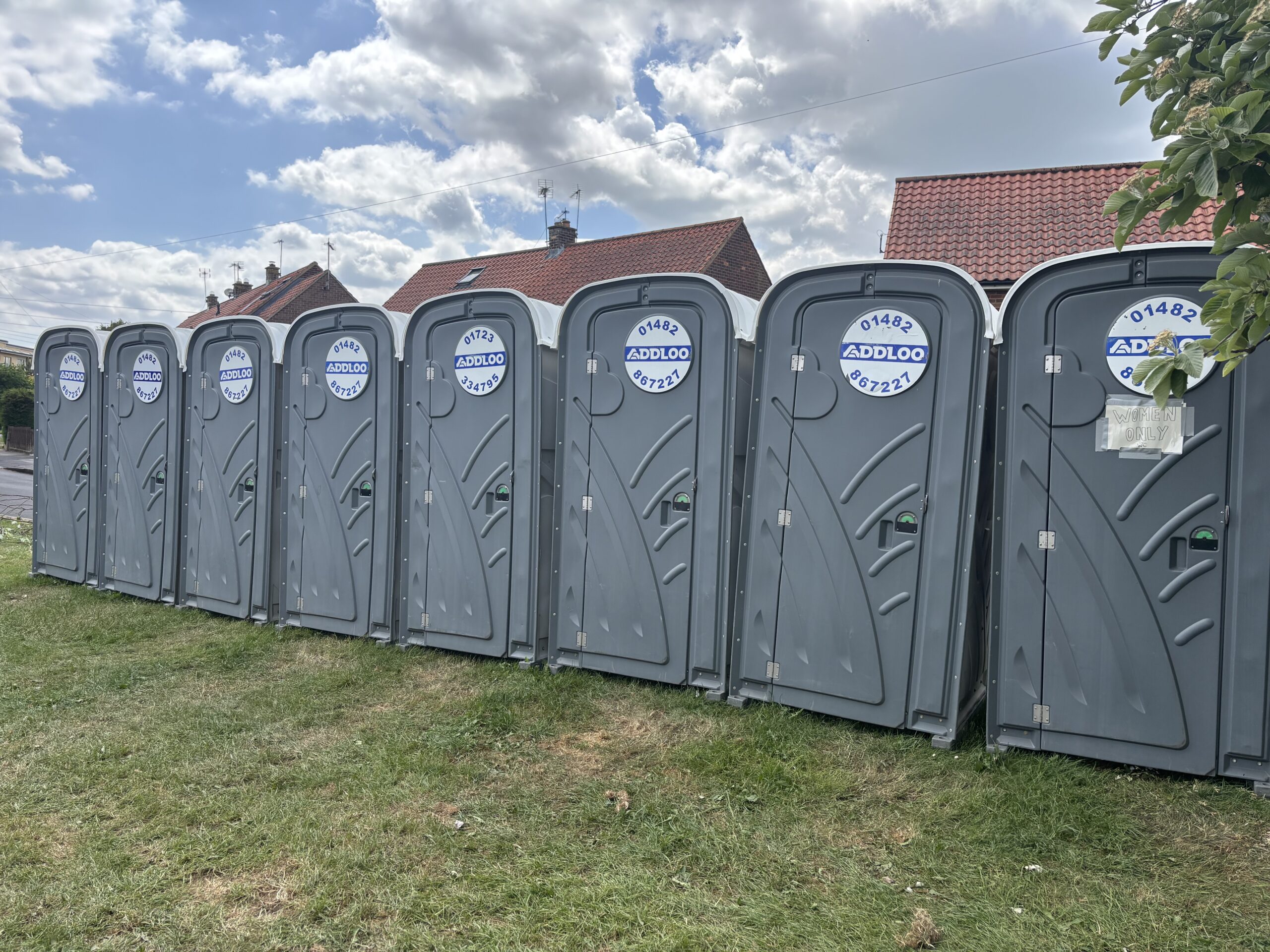 A close-up view of a row of grey portable toilets situated on grass. The toilet on the far right has a handwritten paper sign taped to the door that reads "WOMEN ONLY." The tops of brick houses and leafy trees are visible in the background.