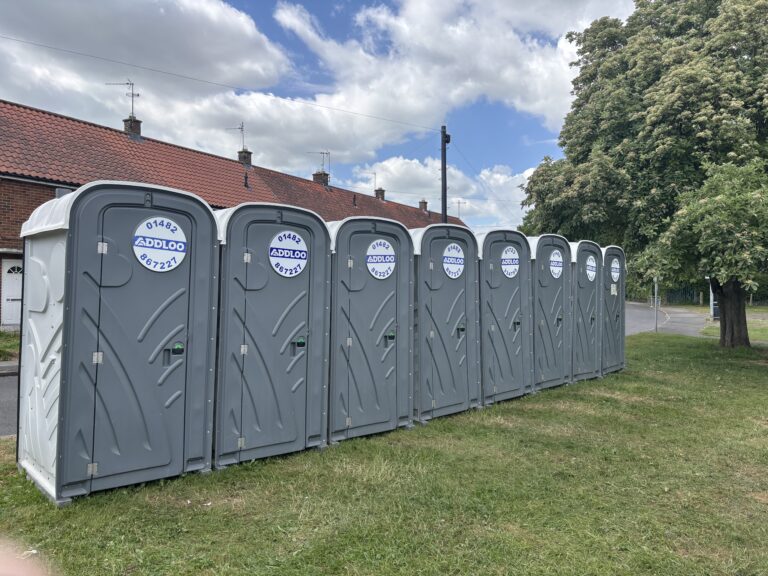 A row of eight identical grey portable toilets with white roofs lined up on a grassy area. Each door features a circular blue and white sticker with the text "ADDLOO" and a phone number. In the background, there are brick residential houses with red roofs under a cloudy blue sky.