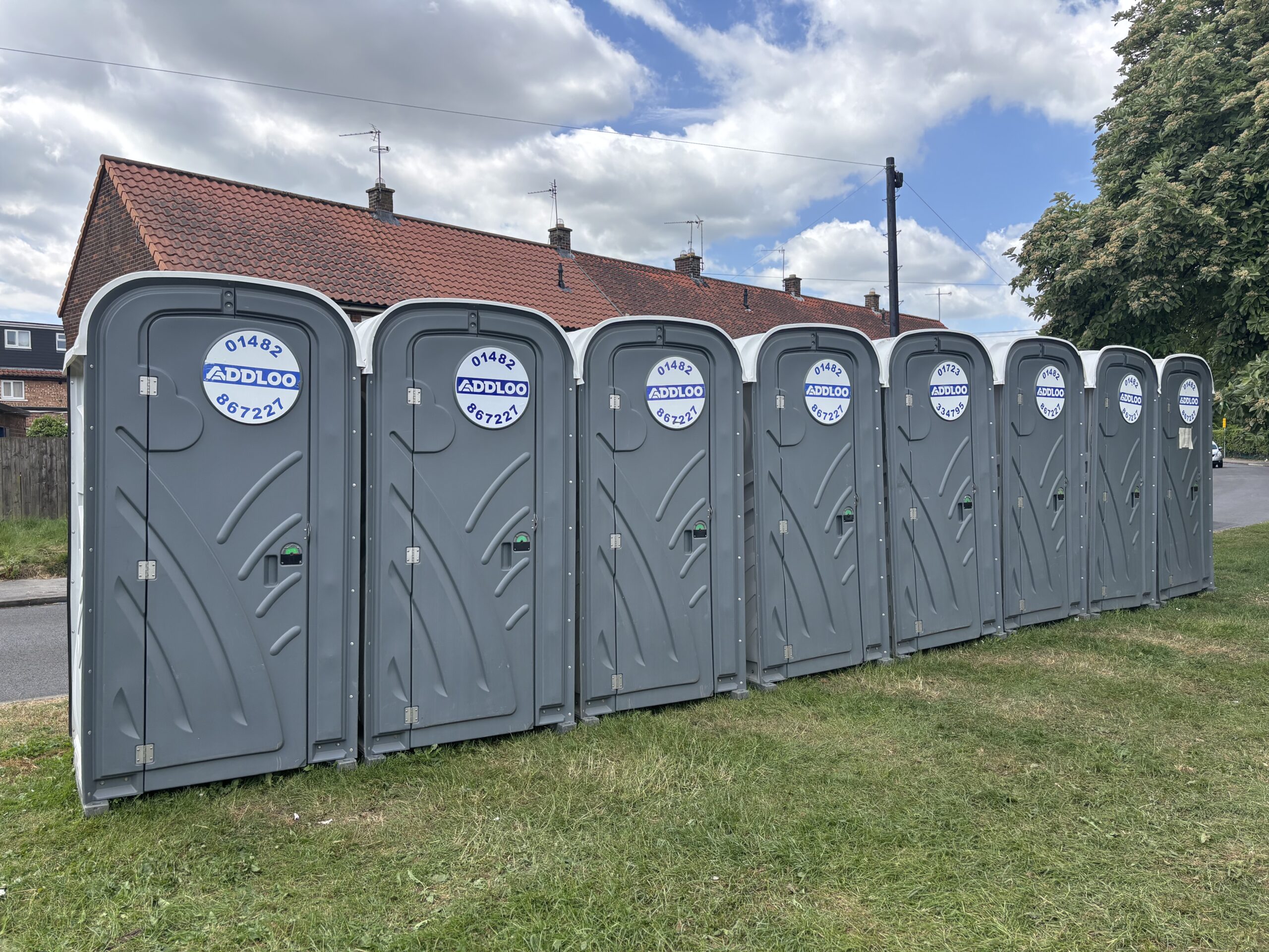 A perspective shot of a line of grey portable toilets with "ADDLOO" branding. The units are positioned on a green lawn in front of a row of brick terraced houses with red tiled roofs and chimneys.