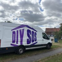 A white Ford transit van parked on a grassy field. The side of the van features a large, stylised purple logo that reads "DIY SOS," designed to look like the frame of a house. The sky above is filled with white and grey clouds.