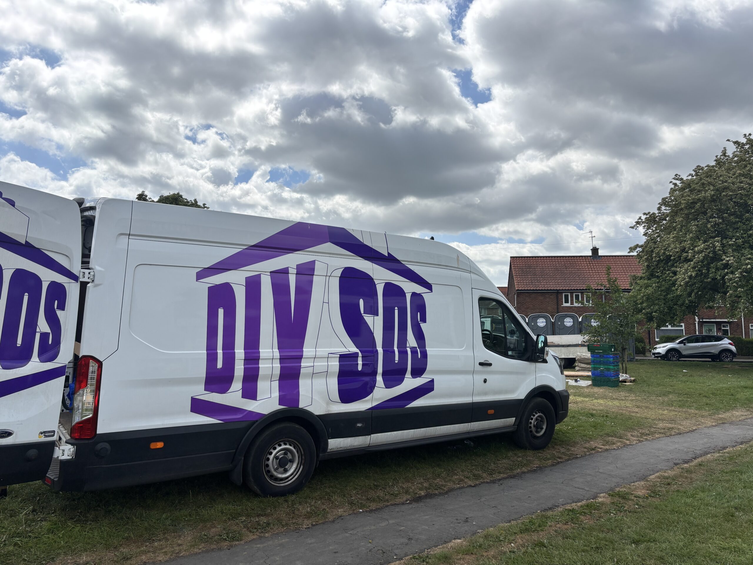 A white Ford transit van parked on a grassy field. The side of the van features a large, stylised purple logo that reads "DIY SOS," designed to look like the frame of a house. The sky above is filled with white and grey clouds.