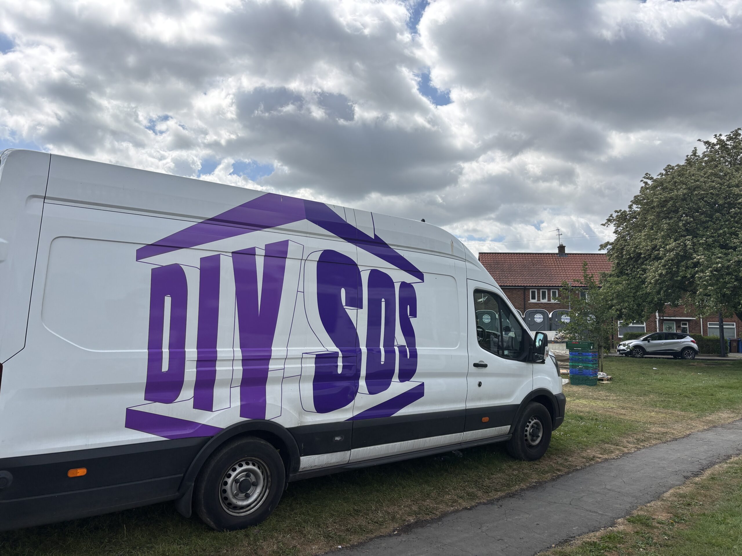 A side view of a white van parked on grass, displaying the large purple text "DIY SOS" inside a house-shaped graphic. In the background, there is a red-brick building and a few stacked crates near a tree.