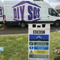 A white van with purple "DIY SOS" branding parked behind a construction site safety sign. The sign features the BBC logo and reads "DIY SOS the big build" followed by "SITE SAFETY STARTS HERE!" and a list of safety warnings and muster point information.