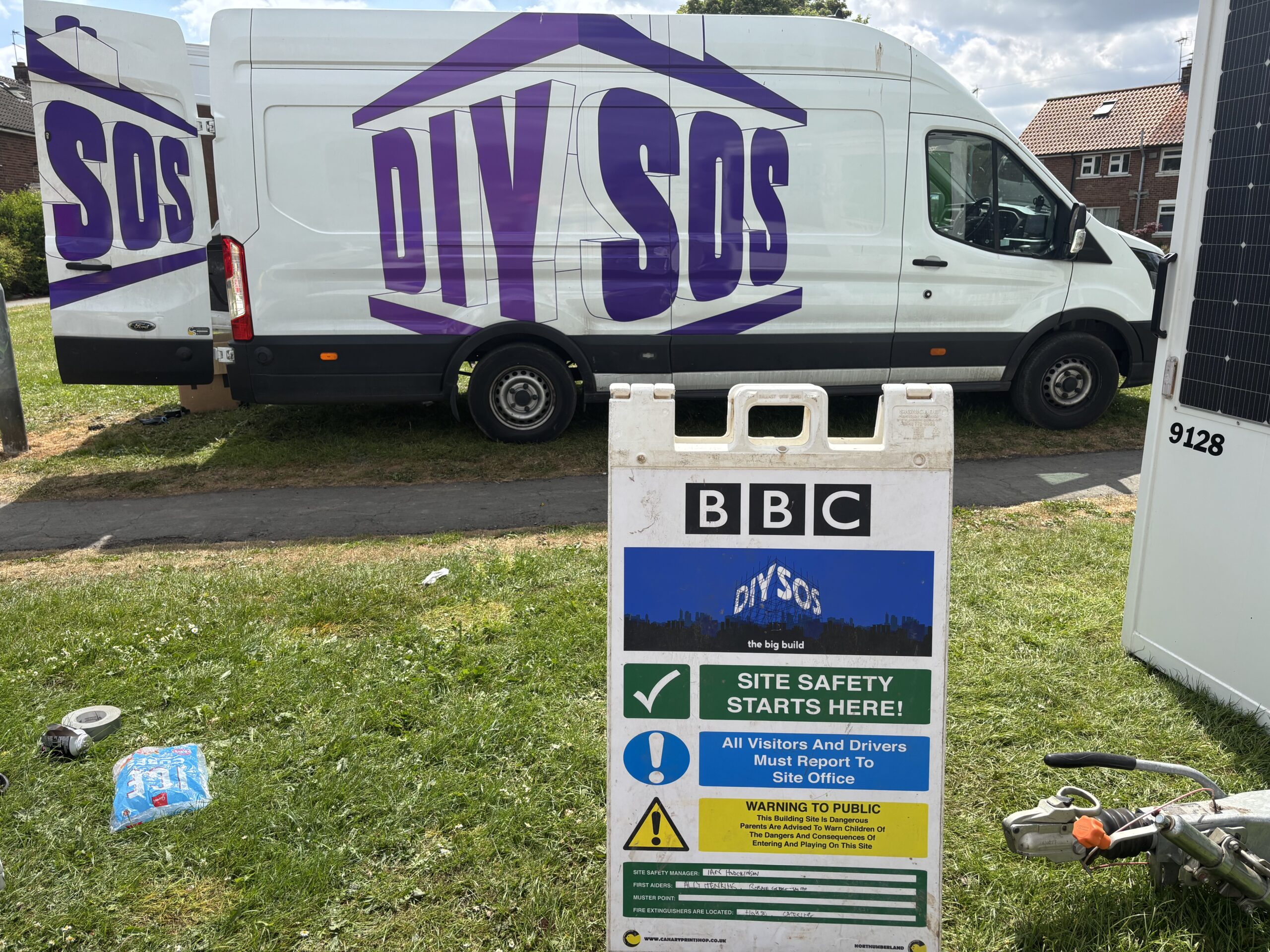 A white van with purple "DIY SOS" branding parked behind a construction site safety sign. The sign features the BBC logo and reads "DIY SOS the big build" followed by "SITE SAFETY STARTS HERE!" and a list of safety warnings and muster point information.