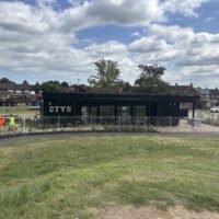 A distant view of a black modular building with "CTYH" lettering, enclosed by a white metal fence. A worker in an orange high-visibility vest sits on a bench to the left. Behind the building, a large flatbed truck carrying construction materials is parked on the street in front of brick houses.