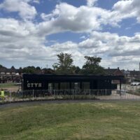A wide landscape shot of a long black building labeled "CTYH" situated in a park. A white metal fence surrounds the building's perimeter. Workers in high-visibility vests are visible near the building and sitting on a bench. A large grassy slope fills the foreground, with residential houses and trees in the distance.
