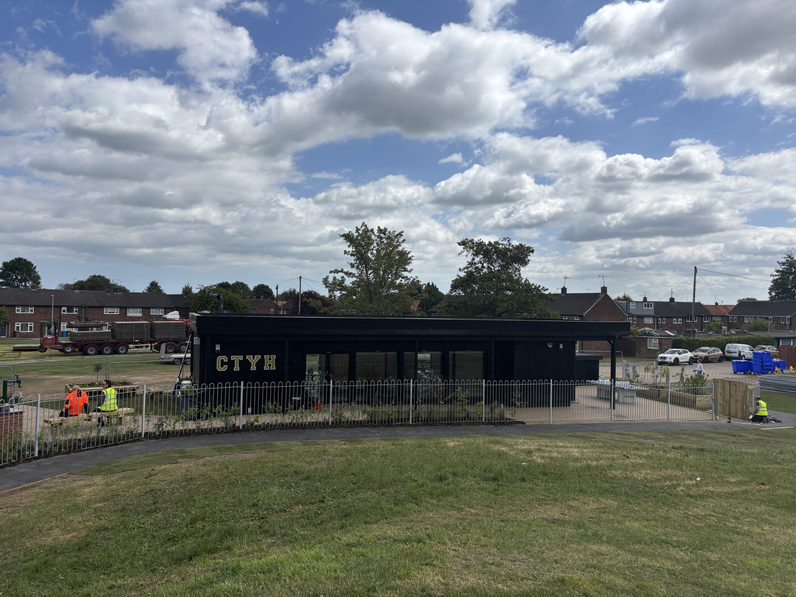A wide landscape shot of a long black building labeled "CTYH" situated in a park. A white metal fence surrounds the building's perimeter. Workers in high-visibility vests are visible near the building and sitting on a bench. A large grassy slope fills the foreground, with residential houses and trees in the distance.