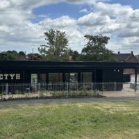 A straight-on view of a rectangular black building featuring large glass windows and gold "CTYH" lettering on the facade. A white metal fence runs across the front, separating the building from a grassy slope in the foreground. Trees and rooftops are visible behind the structure under a sky filled with white clouds.
