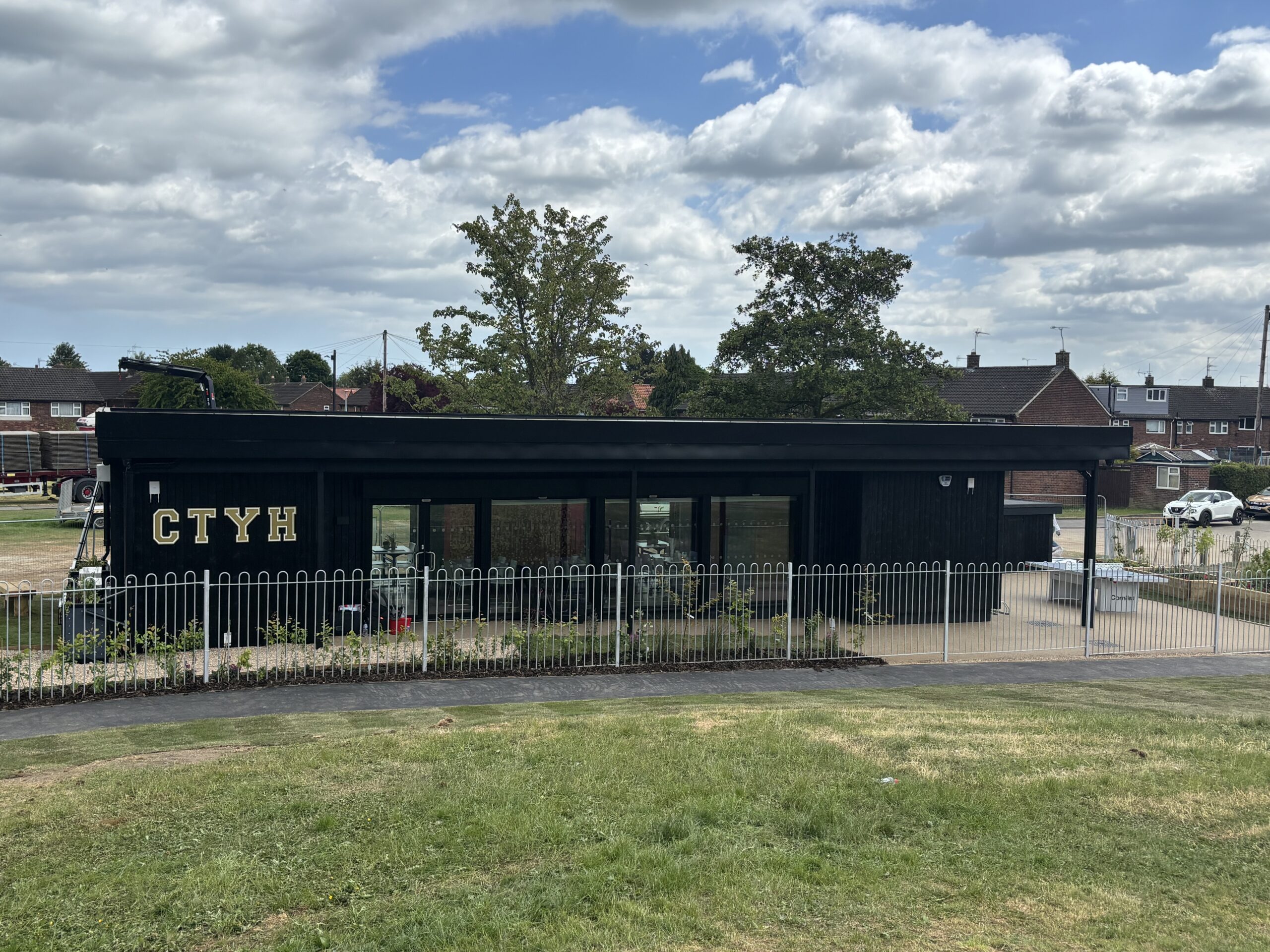 A straight-on view of a rectangular black building featuring large glass windows and gold "CTYH" lettering on the facade. A white metal fence runs across the front, separating the building from a grassy slope in the foreground. Trees and rooftops are visible behind the structure under a sky filled with white clouds.
