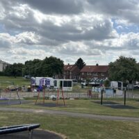 A view of a park playground with a blue circular spinner and swing frames in the foreground. Behind a metal fence, a construction staging area is visible, featuring white vans with purple "DIY SOS" logos, portable toilets, and various building materials. A row of brick residential houses lines the background under a dramatic cloudy sky.