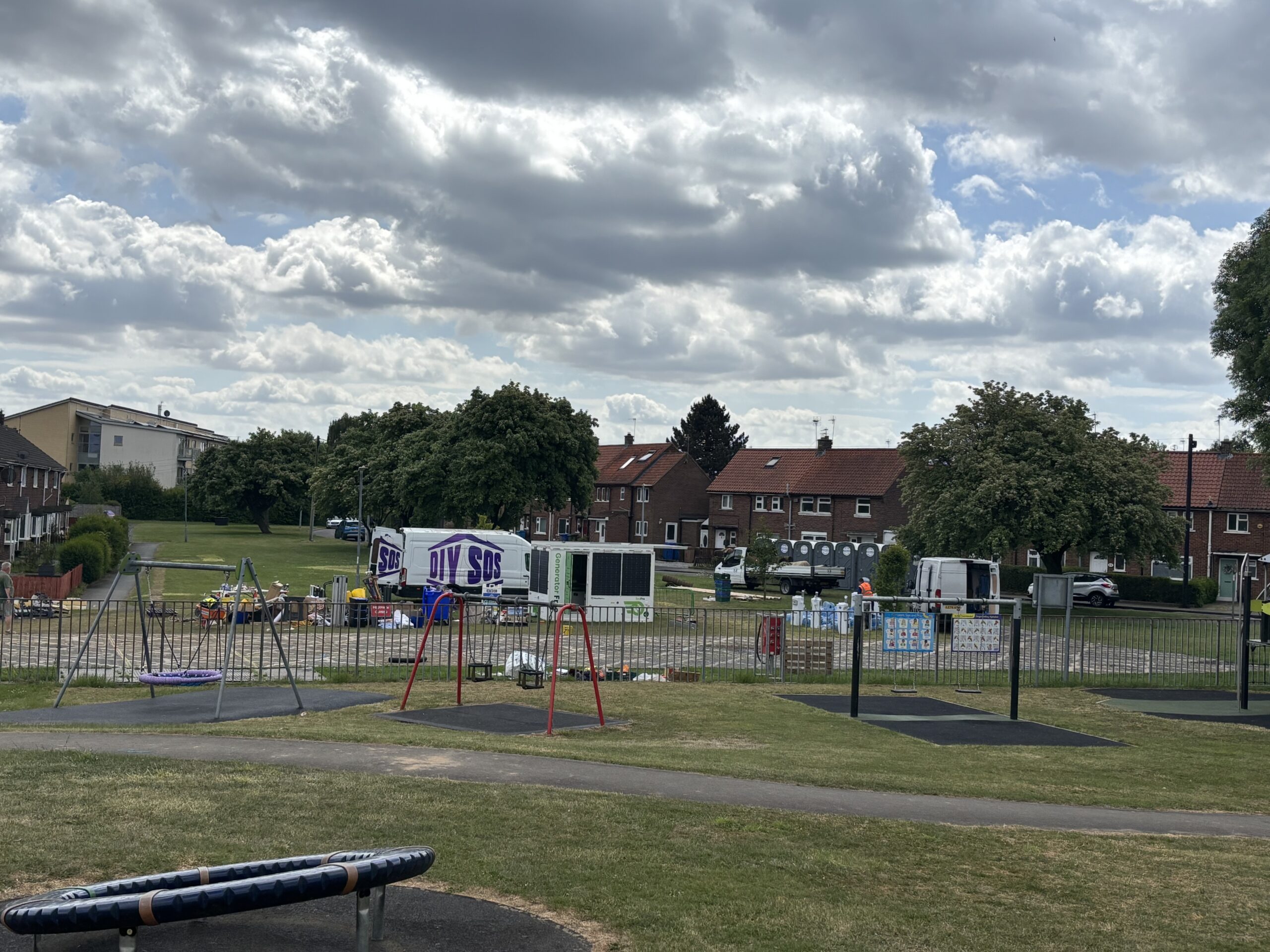 A view of a park playground with a blue circular spinner and swing frames in the foreground. Behind a metal fence, a construction staging area is visible, featuring white vans with purple "DIY SOS" logos, portable toilets, and various building materials. A row of brick residential houses lines the background under a dramatic cloudy sky.