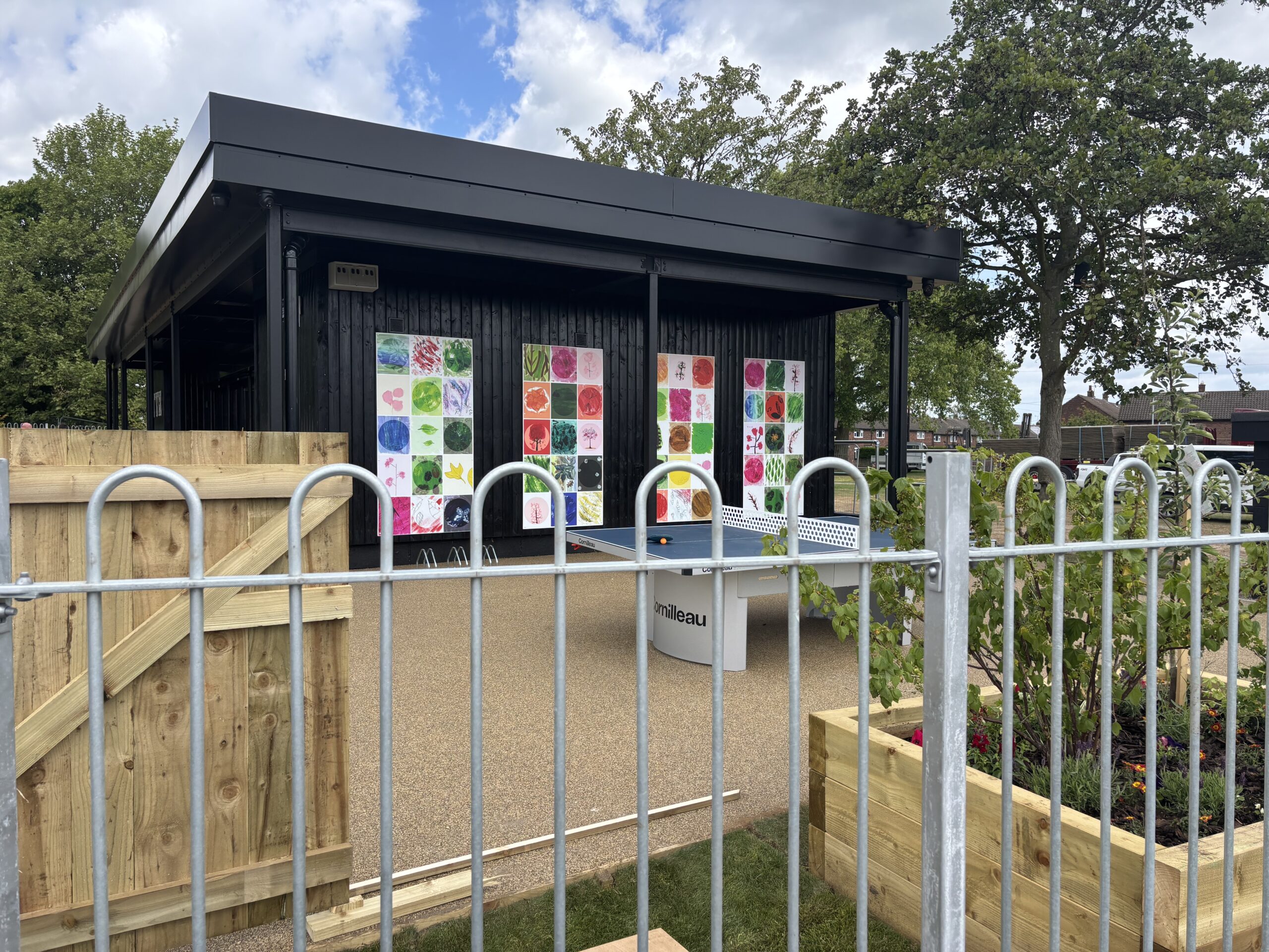 A view looking through a metal and wooden fence at a covered outdoor patio attached to a black building. The building wall is decorated with three vertical panels of colorful, square artwork. A blue table tennis table sits on the paved surface, shaded by the building's overhang.