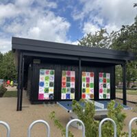 An outdoor recreational space featuring a blue table tennis table in front of a black building wall adorned with four vertical panels of colorful artwork. White metal hoop barriers are in the immediate foreground. To the left, two people in high-visibility vests are seen sitting on a bench in the distance.