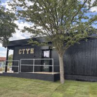 A corner view of a modern, single-story building with black timber cladding. The gold letters "CTYH" are mounted on the exterior wall. A metal ramp leads to a glass entrance door. A large tree with green leaves stands in the foreground, partially obscuring the building.