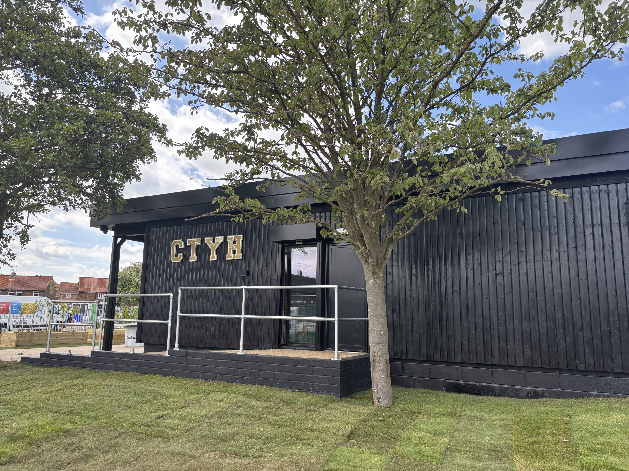 A corner view of a modern, single-story building with black timber cladding. The gold letters "CTYH" are mounted on the exterior wall. A metal ramp leads to a glass entrance door. A large tree with green leaves stands in the foreground, partially obscuring the building.